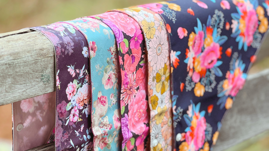 A set of six sun-safe neck scarves with different floral patterns draped over a weathered wooden fence outdoors.